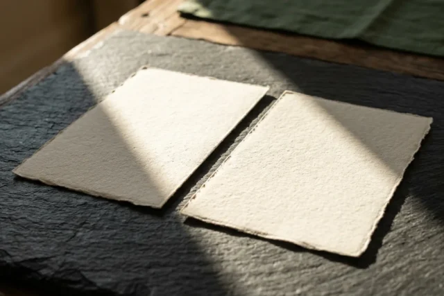 A thick, textured blank paper document cleanly sliced in two on a dark slate tabletop, the two halves slightly separated under a sharp, dramatic shaft of morning light that casts long, geometric shado